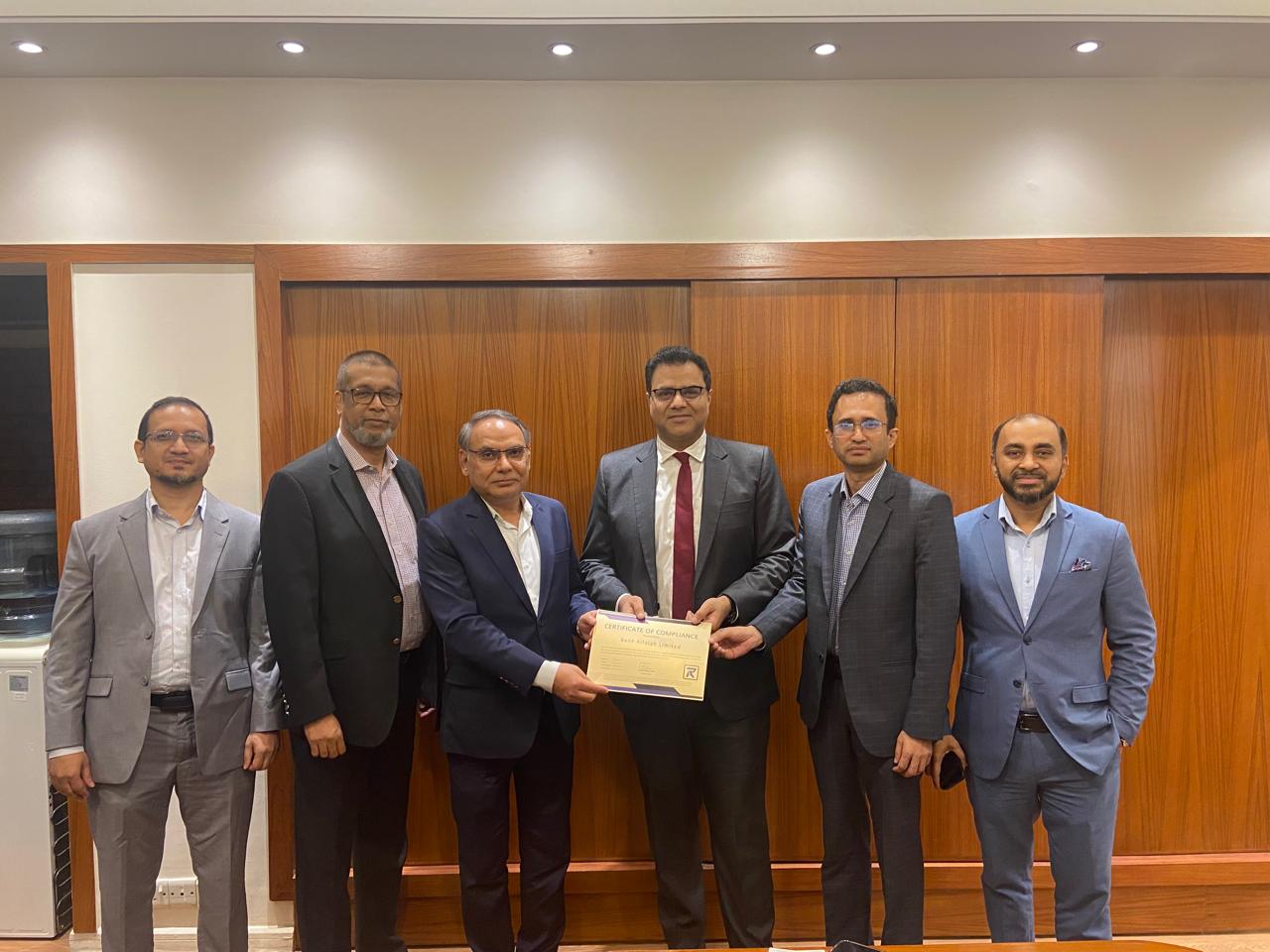 A group of six professionally dressed men stand together, with two of them holding a certificate of compliance featuring the Risk Associates logo. The setting is a formal indoor space with a wooden backdrop, reflecting a professional achievement or recognition event related to governance, risk, and compliance
